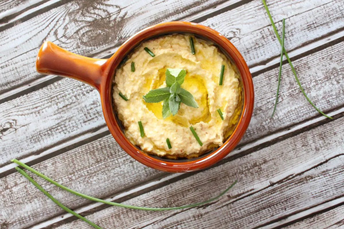 Cauliflower hummus in a small bowl with a handle. The hummus is garnished with a sprig of mint in the middle, and some diced chives around the edges. The bowl in which the cauliflower hummus sits, has a brown rim. The bowl sits on a table, and is surrounded by 2 long pieces of chives, on both the top right corner, and the bottom left corner.