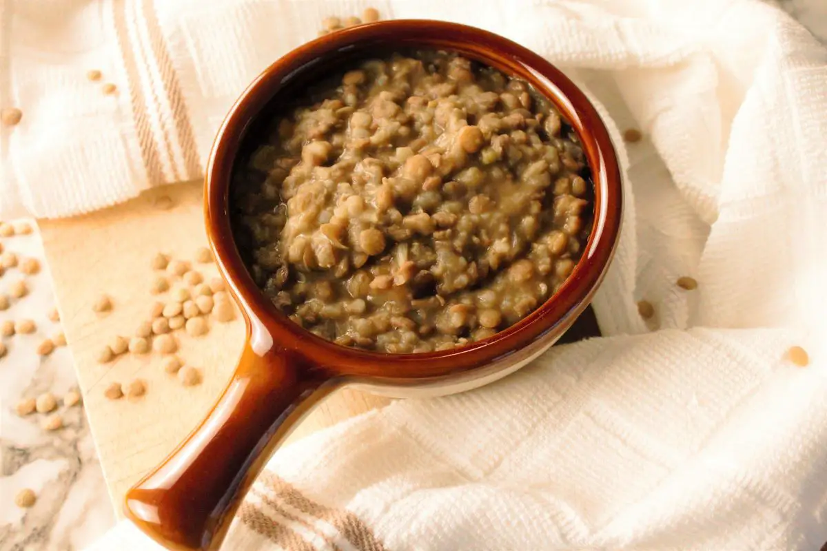 Lentil Soup in a small bowl with a handle. The handle and the rim of the bowl are of a brown color. There is a white kitchen towel with beige stripes on its ends that's curving around the bowl over to the right corner. The lentil soup bowl is sitting over a cutting board. Grains of lentils are sprinkled over the board, and around the bowl.