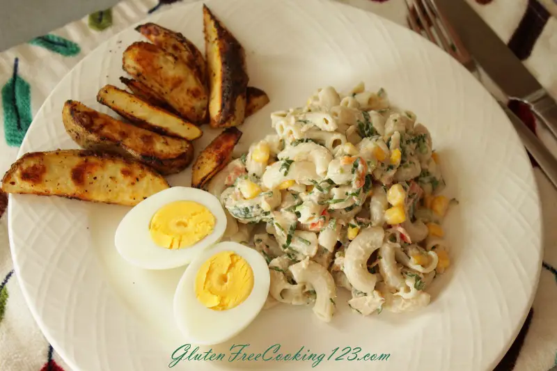 Pasta salad in a white serving plate. an egg sliced in half length wise is at the bottom left corner of the pasta salad, and some oven baked fries are at the top left corner of the pasta salad.