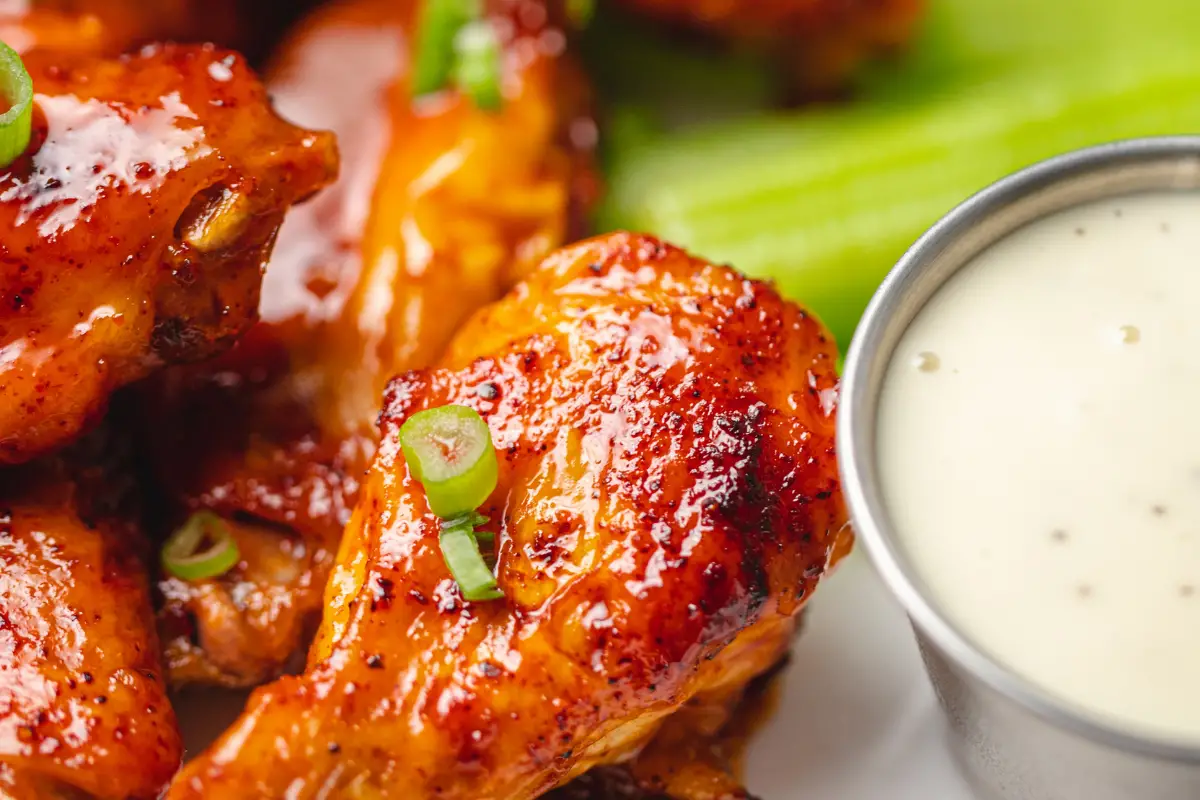 Baked buffalo wings sprinkled with spring onions. A bowl of blue cheese dipping sauce sits to the bottom right corner of the wings. A few green stems of celery sit to the top of the sauce and under the wings.