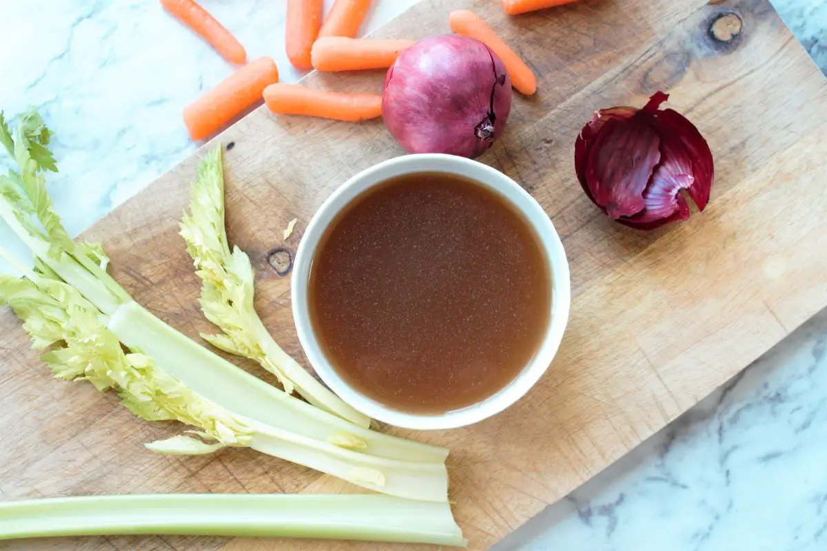 Bone broth in a white bowl over a cutting board. Surrounding the bowl starting from the left side to the right, are, 3 celery stalks, baby carrots, a red onion, and a red onion's outer peel.