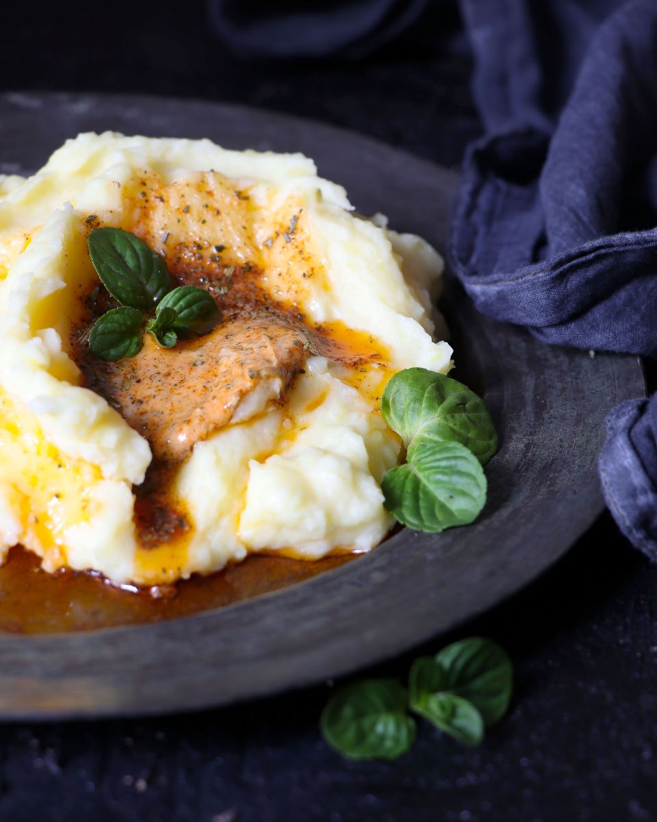 Creamy mashed potatoes in a dark plate with gravy drizzled over it, and garnished with oregano.