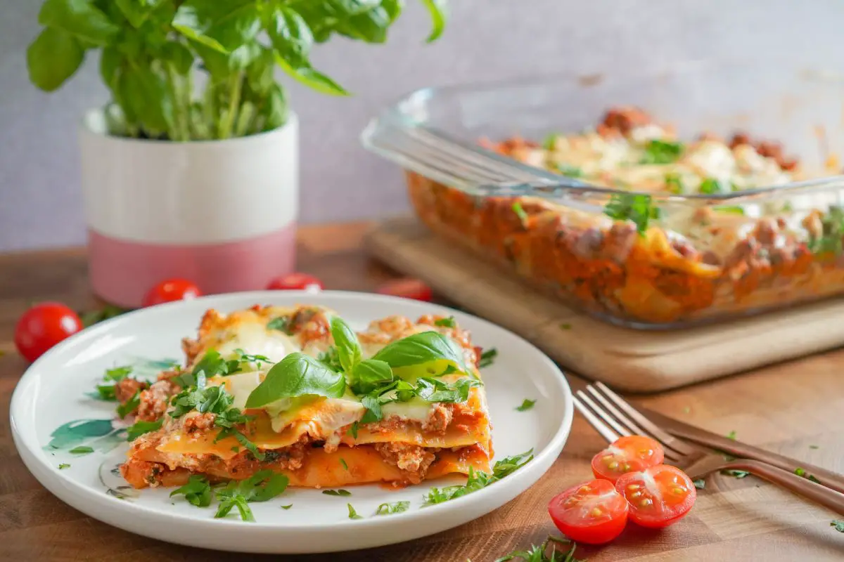 A slice of lasagna on a serving plate on the lower left corner. The plate is surrounded with sliced cherry tomatoes. A glass baking dish full of lasagna sits at the top right of the picture, and a small cylinder vase filled with basil sits at the top left of the picture.