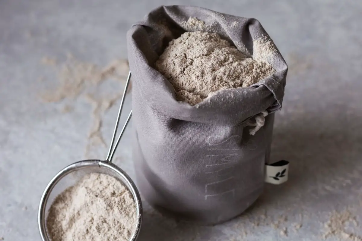 Rice flour in a fabric bag on a counter surface. Some rice flour in a sieve on the counter, sits beside the bag of flour at the bottom left corner. Some flour is also sprinkle on the counter.